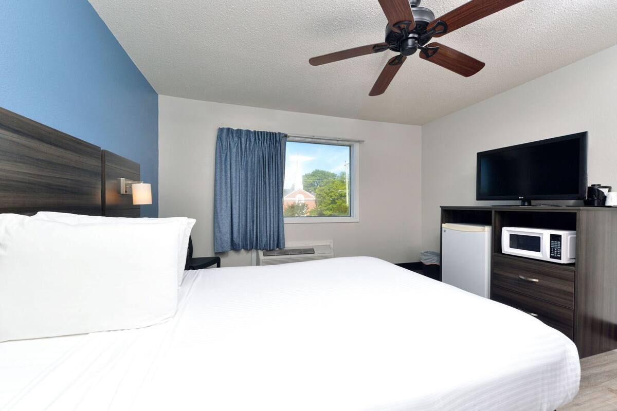 A hotel room with a bed, a ceiling fan, a television, a microwave, and a window with curtains.
