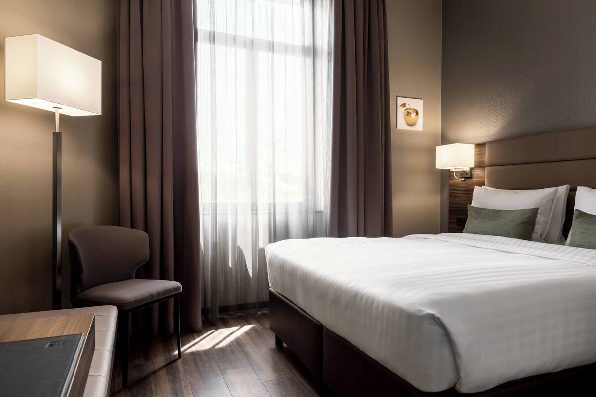 A hotel room with a large bed, a desk, a chair, a window with curtains, and a wall-mounted lamp.