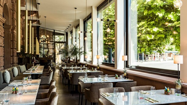 A restaurant with long tables, cushioned chairs, and hanging pendant lights. - AC Hotel Mainz (Mainz)