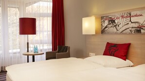 Habitación de hotel con una cama grande, una mesita con dos botellas, una silla y una cortina roja.