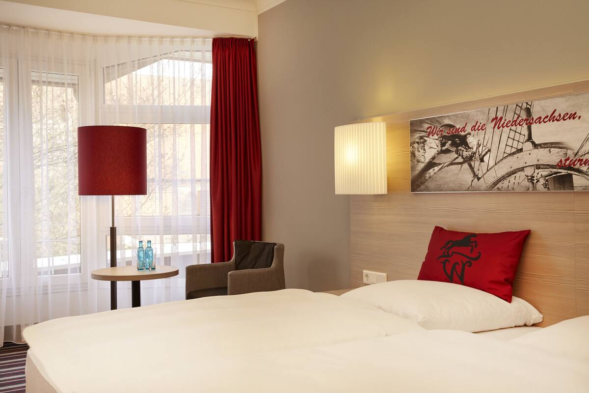 A hotel room with a large bed, a small table with two bottles, a chair, and a red curtain.