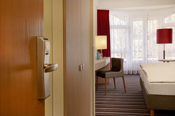 Una habitación de hotel con cama, escritorio, silla, lámpara y ventana con cortinas.