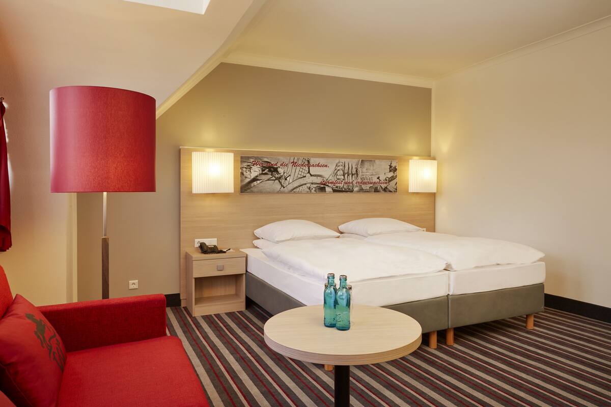 A hotel room with a bed, a red sofa, a small table with bottles, and a lamp.