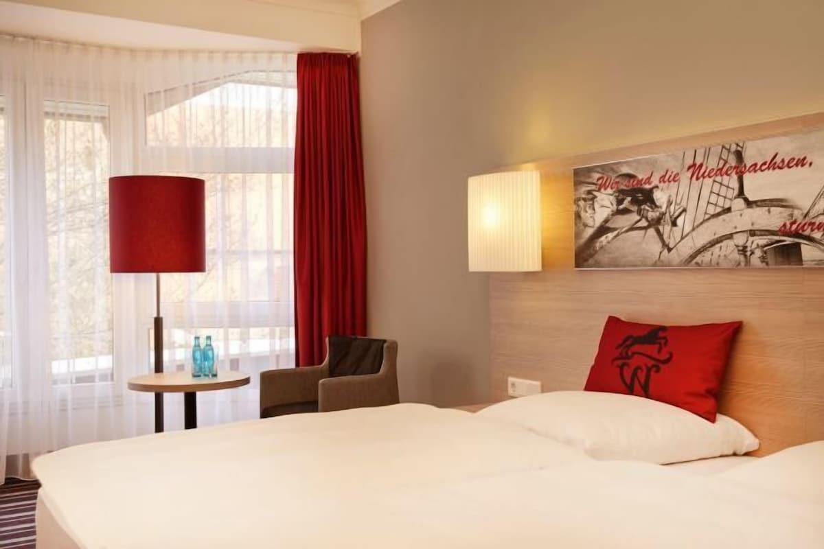 A hotel room with a large bed, a small table with two bottles, a chair, a red curtain, and a wall with a framed picture and text.