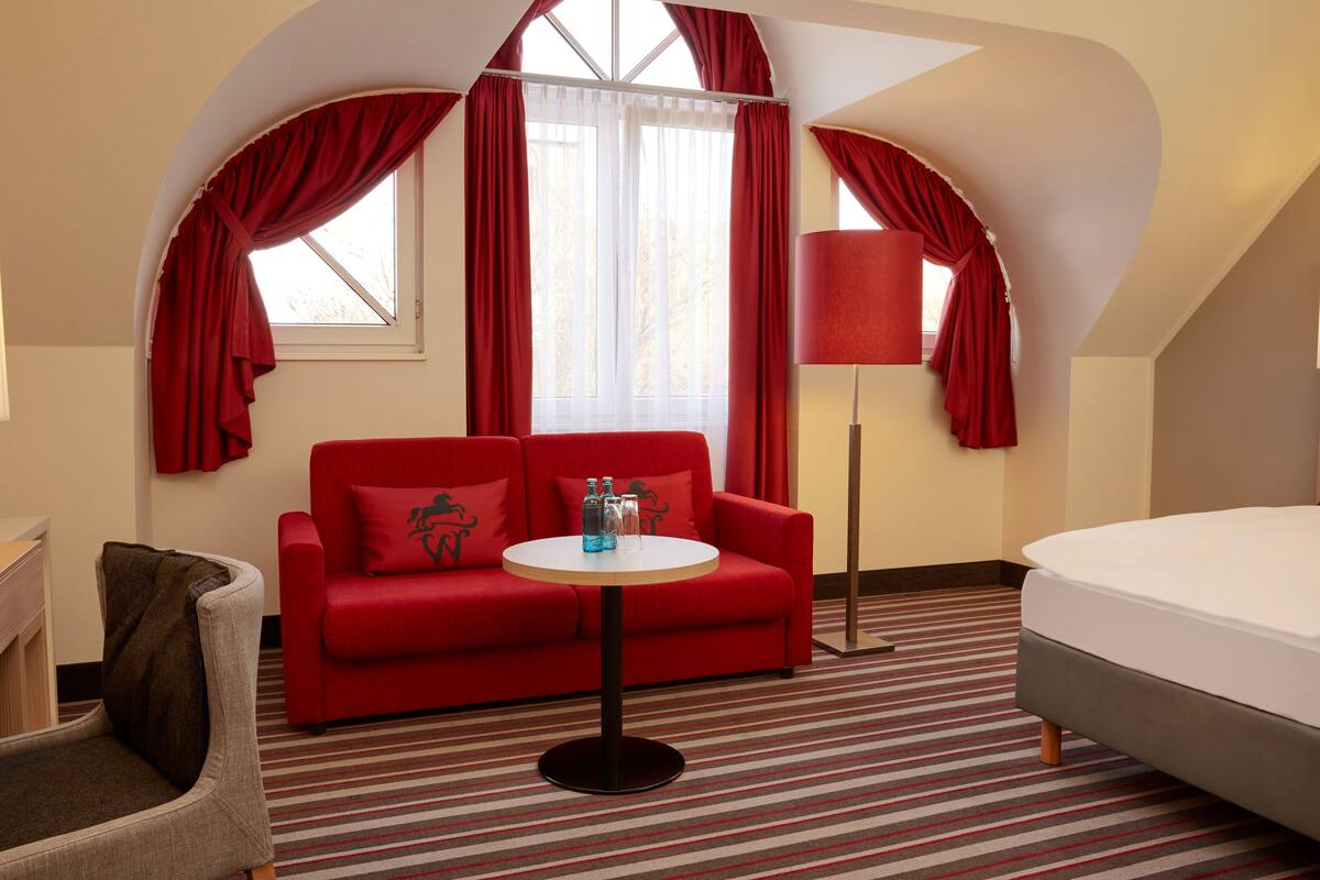 A hotel room with a red sofa, a small round table, a lamp, and a bed.