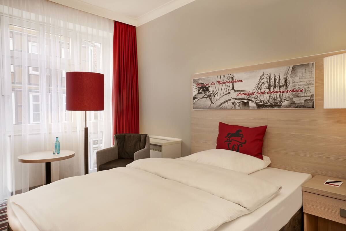 A hotel room with a bed, a nightstand, a chair, a table with a bottle, and a window with curtains.