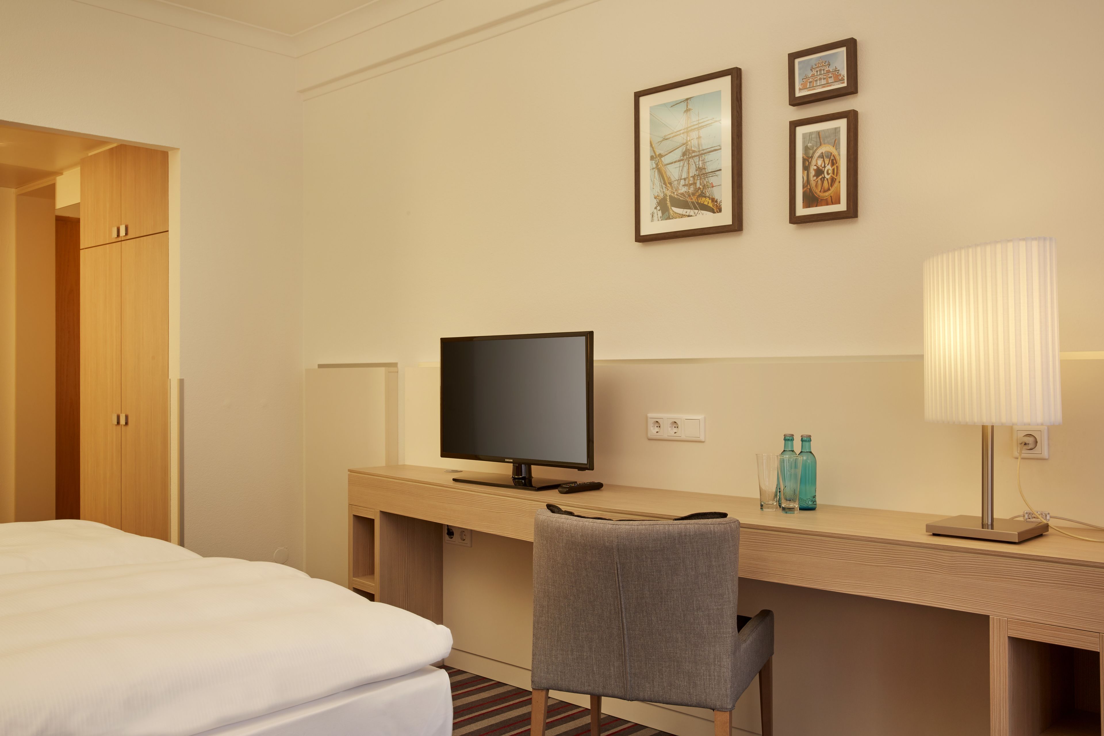 Une chambre d’hôtel avec un lit, un bureau doté d’une télévision, une chaise et une lampe. Des tableaux encadrés ornent le mur.