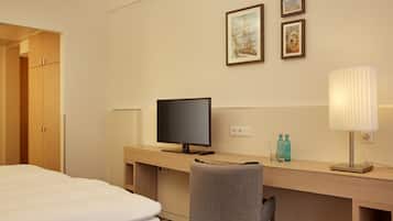A hotel room with a bed, a desk with a television, a chair, and a lamp. There are framed pictures on the wall.