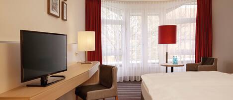 Une chambre d’hôtel avec un lit, un bureau avec une télévision, une chaise, une lampe et une fenêtre avec des rideaux.