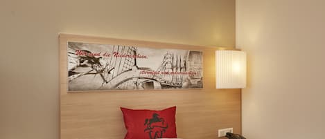 Uma cama bem arrumada com roupa de cama branca, um travesseiro vermelho com silhueta preta, um quadro com uma imagem de um barco sobre uma ponte e um abajur na mesa de cabeceira.