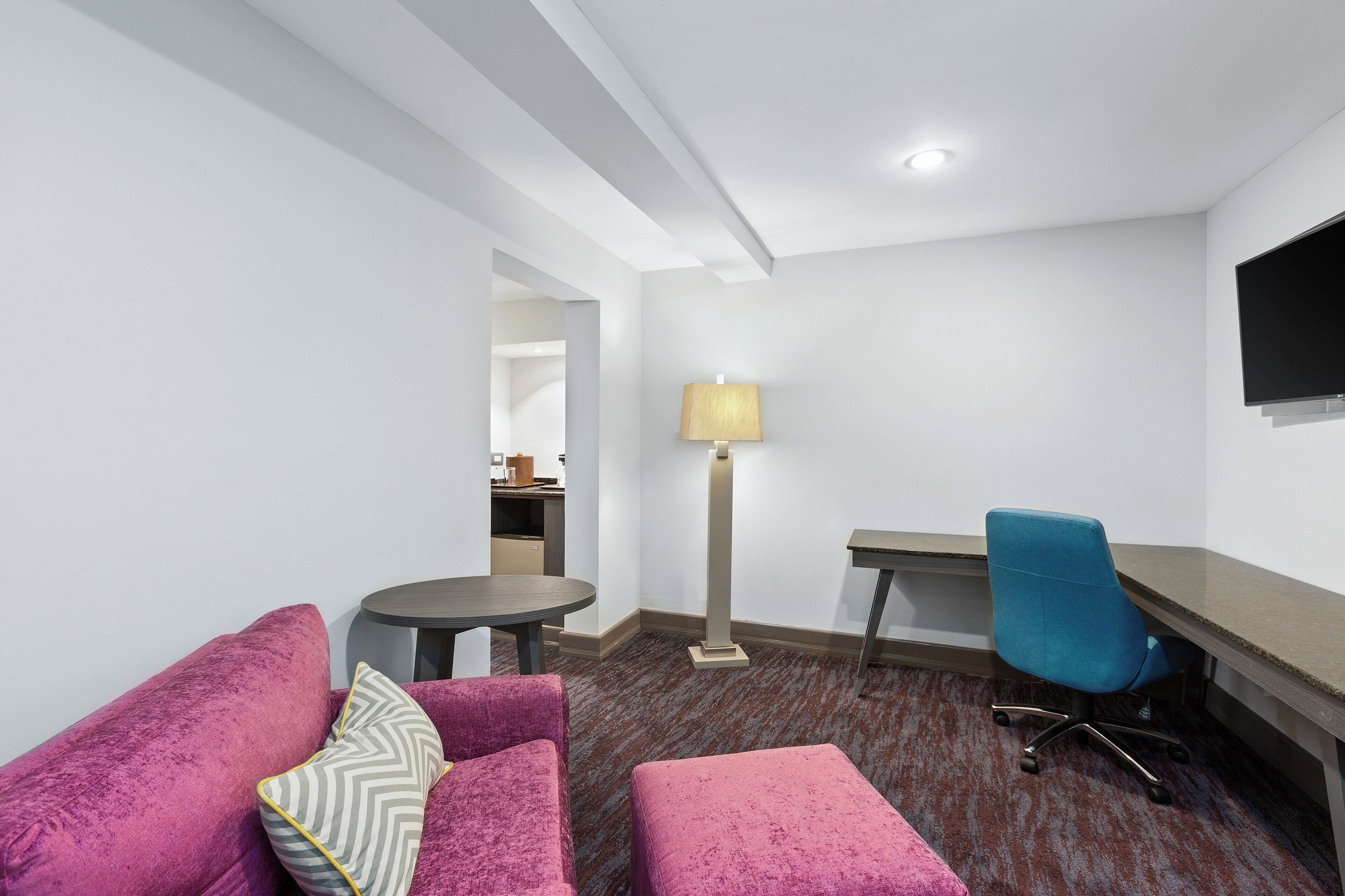 Une chambre d’hôtel moderne avec un canapé rose moelleux, une petite table ronde, un bureau avec une chaise et un téléviseur à écran plat fixé au mur.