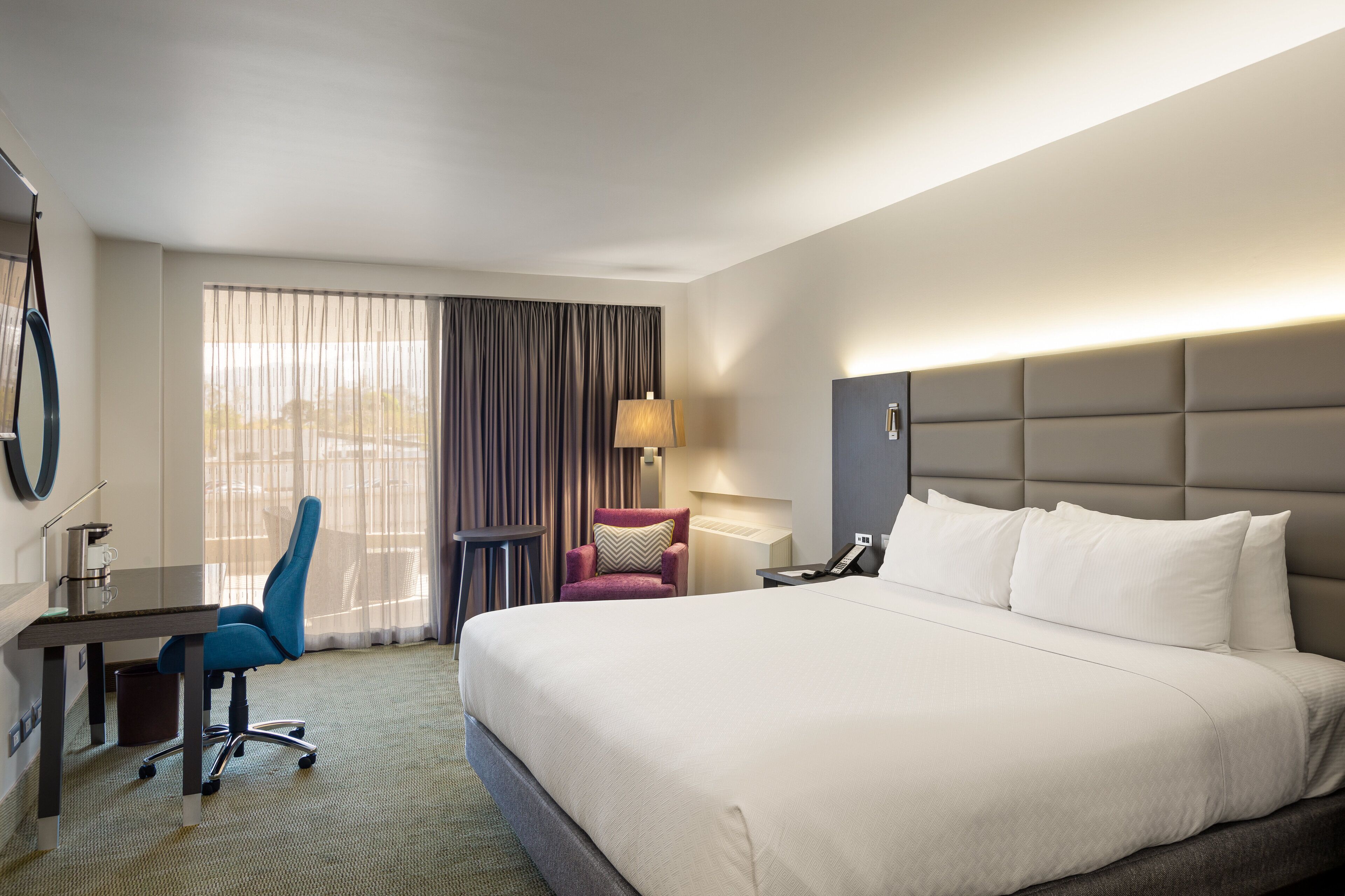 A hotel room with a large bed, a desk with a chair, a small table, and a lamp.