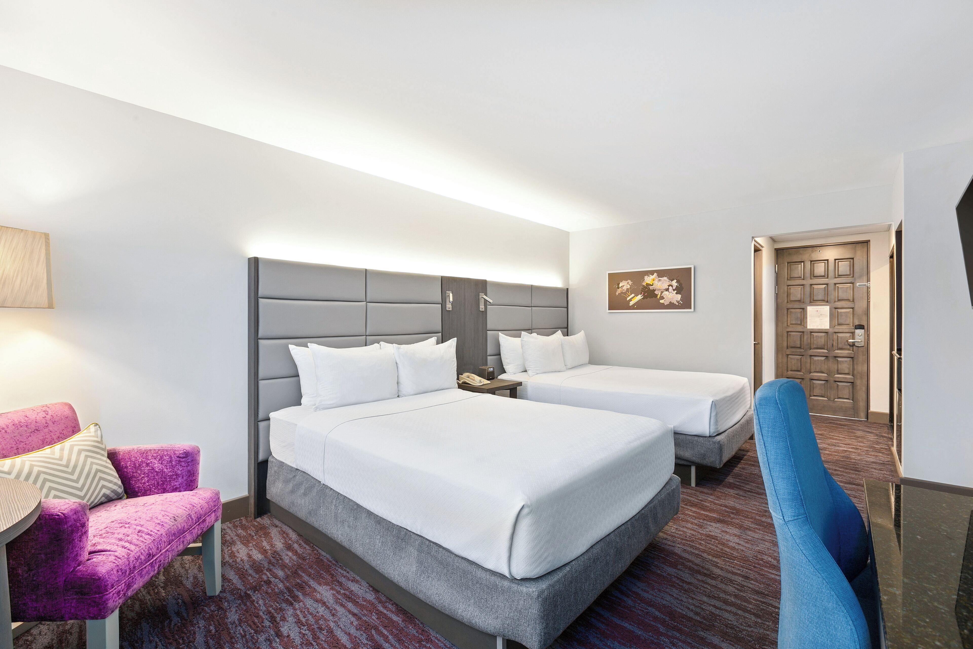 A hotel room with two beds, a chair, a desk, and a door.
