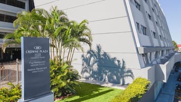 Um edifício moderno com a placa do Crowne Plaza San Jose Corbici Conference Center, apresentando uma área ajardinada com palmeiras e um arbusto bem aparado.
