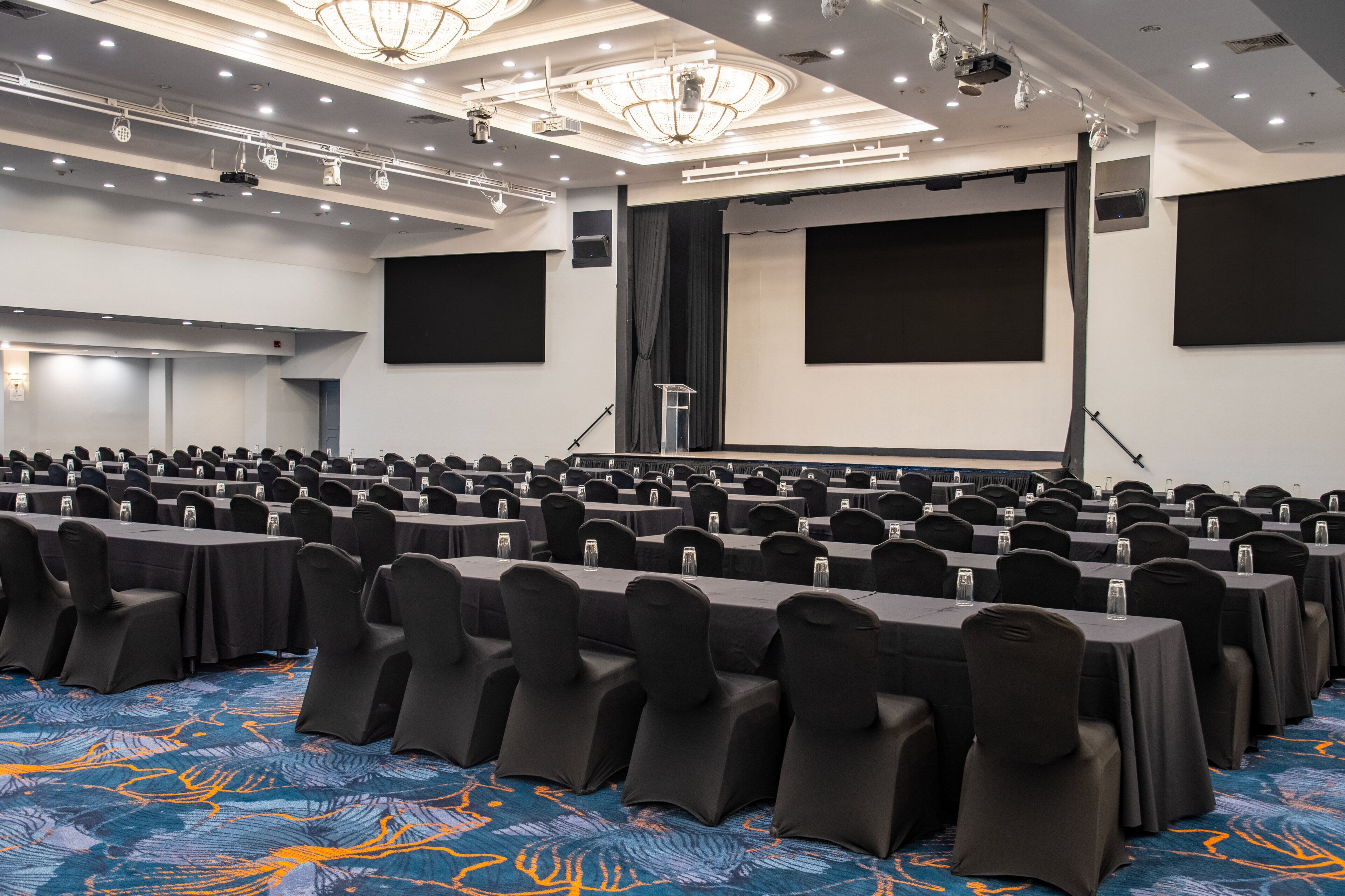 A conference room with rows of tables and chairs, a large screen, and a stage with a podium.