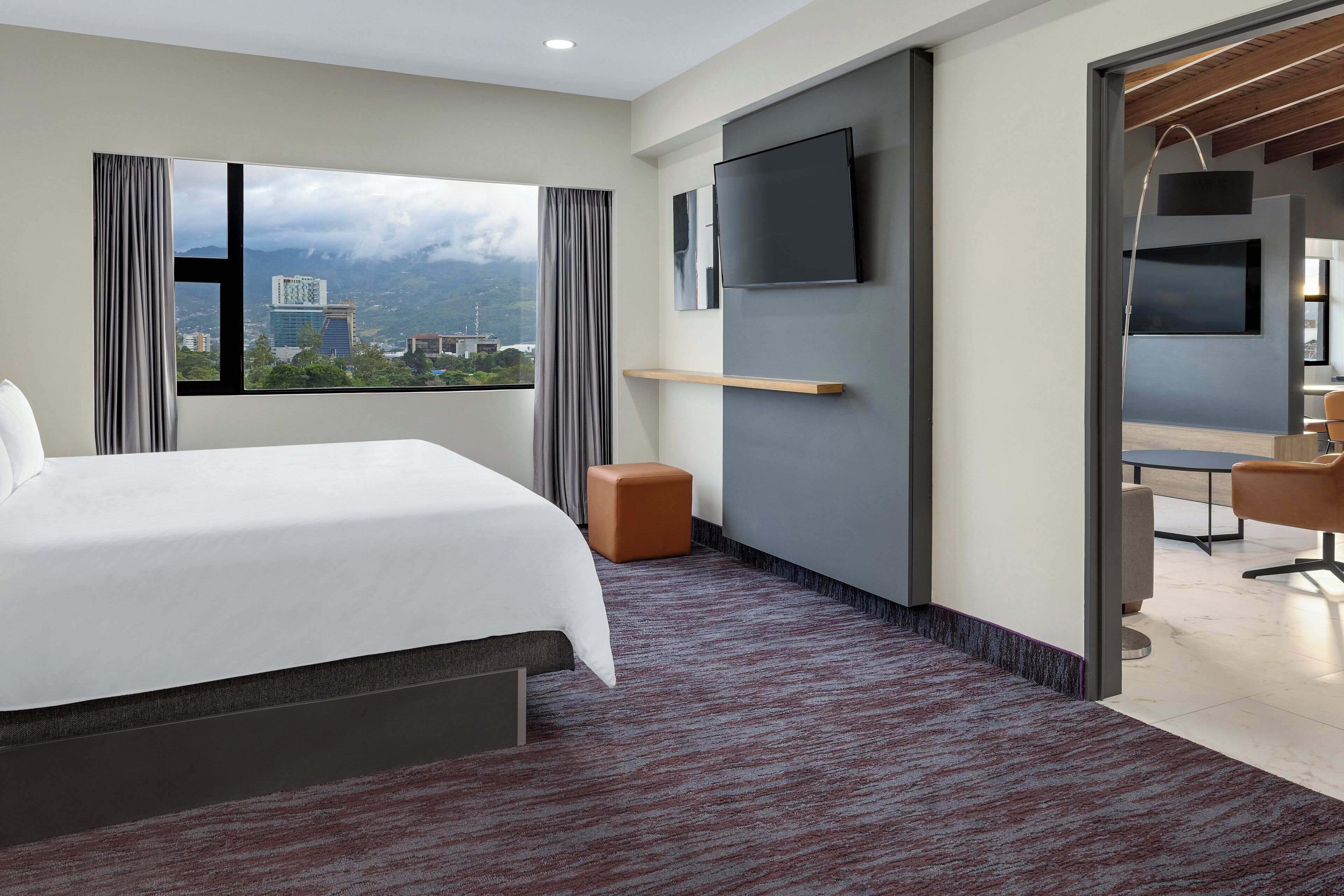 Una habitación de hotel moderna con una cama grande, un televisor montado en la pared y vistas a la ciudad desde la ventana.