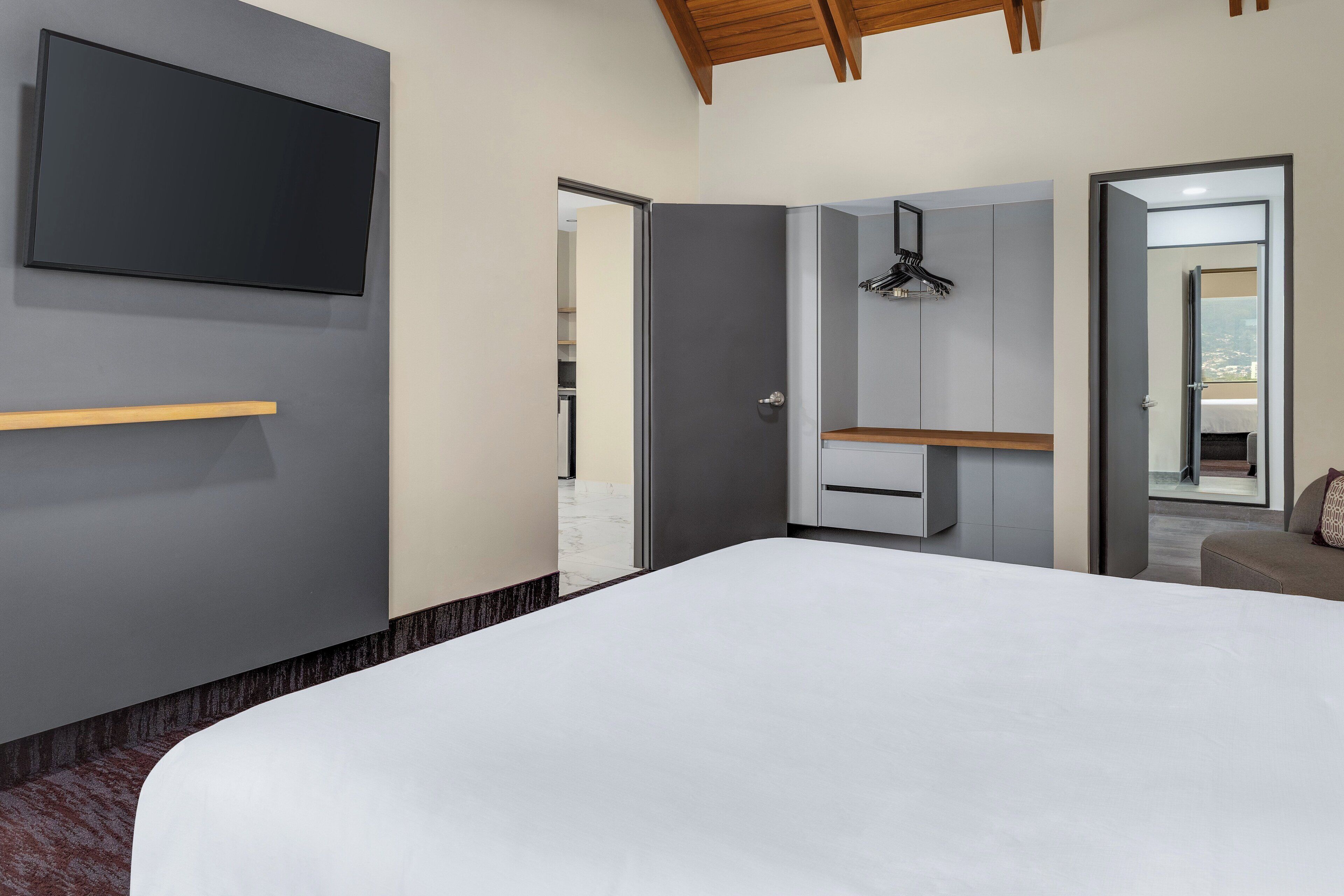 A modern bedroom with a large bed, a TV mounted on the wall, a wardrobe, and a mirror.