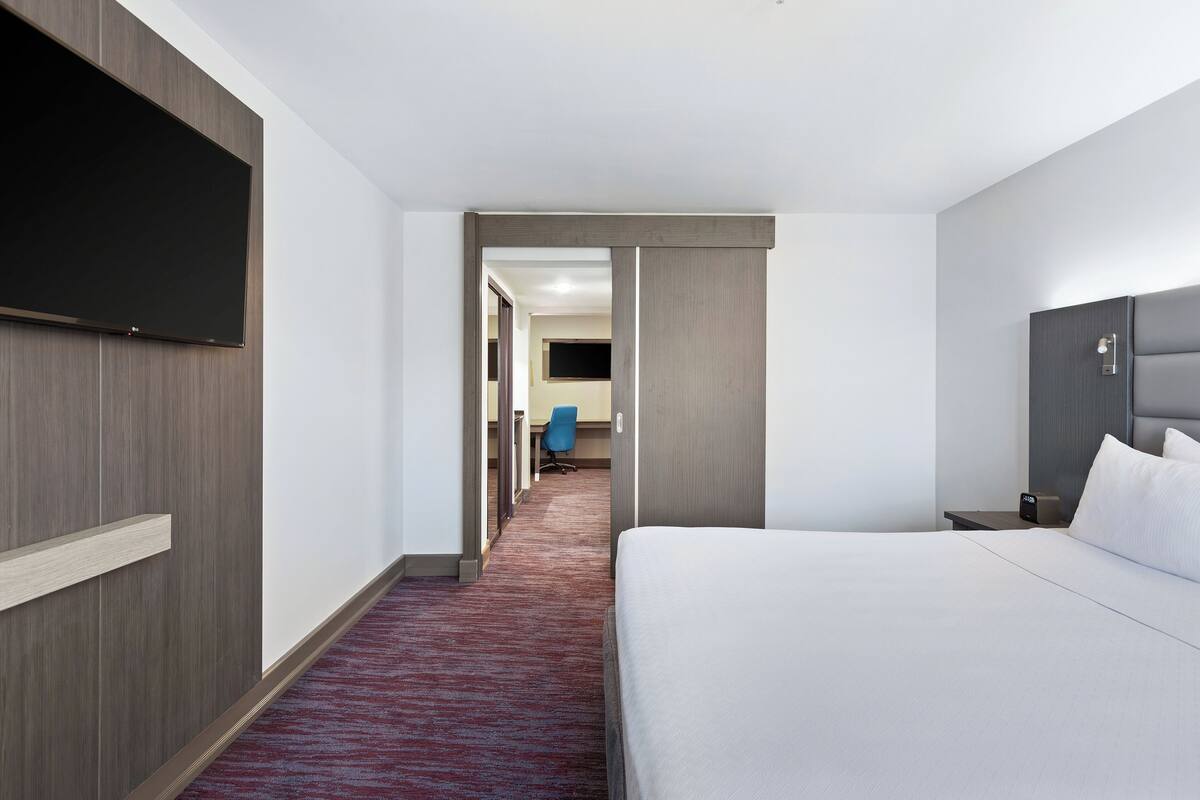 Une chambre d’hôtel avec un grand lit, un téléviseur à écran plat fixé au mur, un bureau avec une chaise et un couloir recouvert d’une moquette rouge.