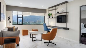 Een moderne woonkamer met een bank, fauteuil en salontafel, een aan de muur bevestigde televisie, en een groot raam met uitzicht op de stad.