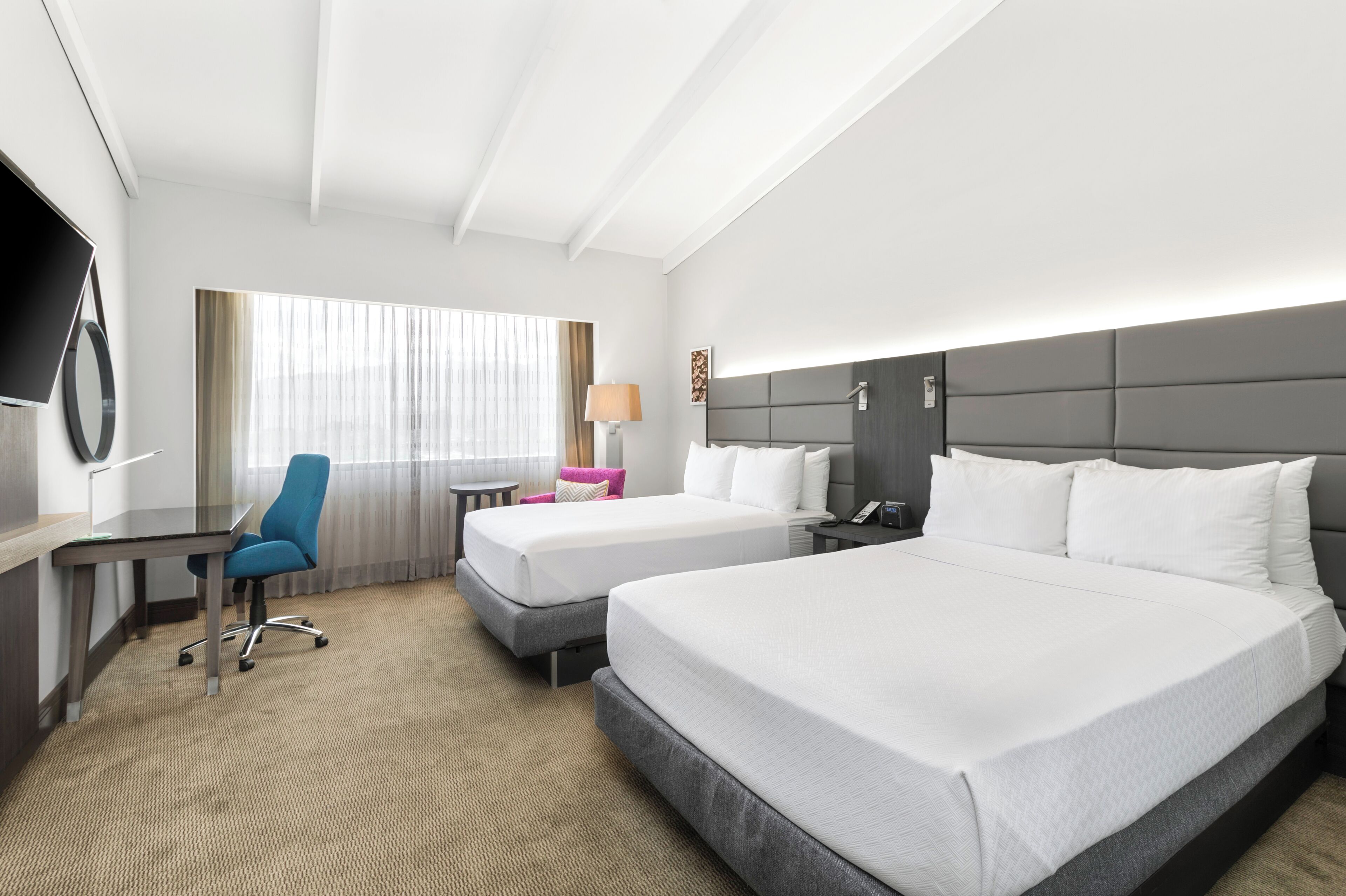 A hotel room with two beds, a desk, a chair, a TV, and a window with curtains.