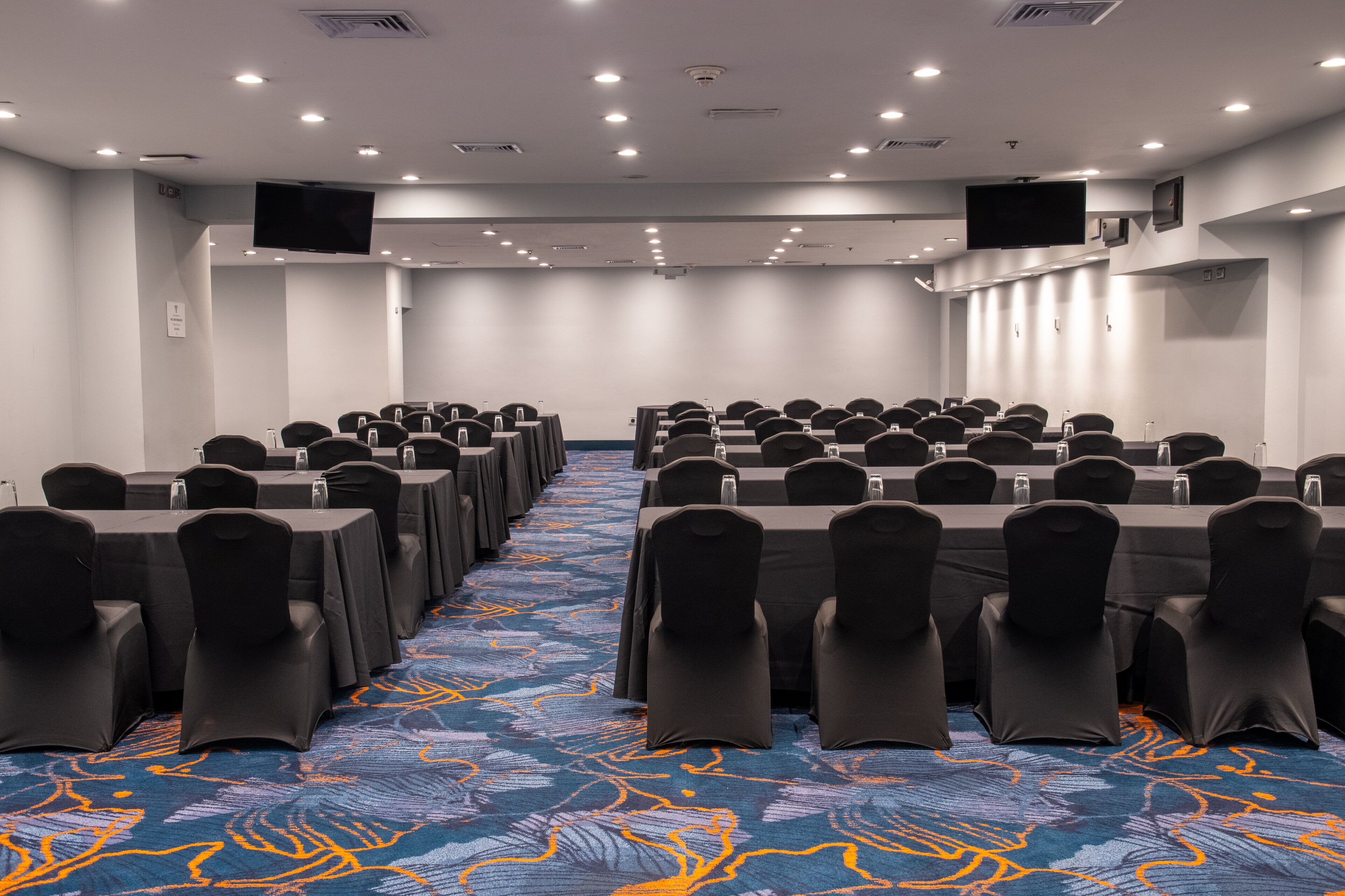Une salle de conférence avec des rangées de tables et de chaises, une moquette à motifs et des luminaires au plafond.