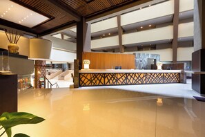 A modern hotel lobby with a central reception desk, a staircase, and a large plant.