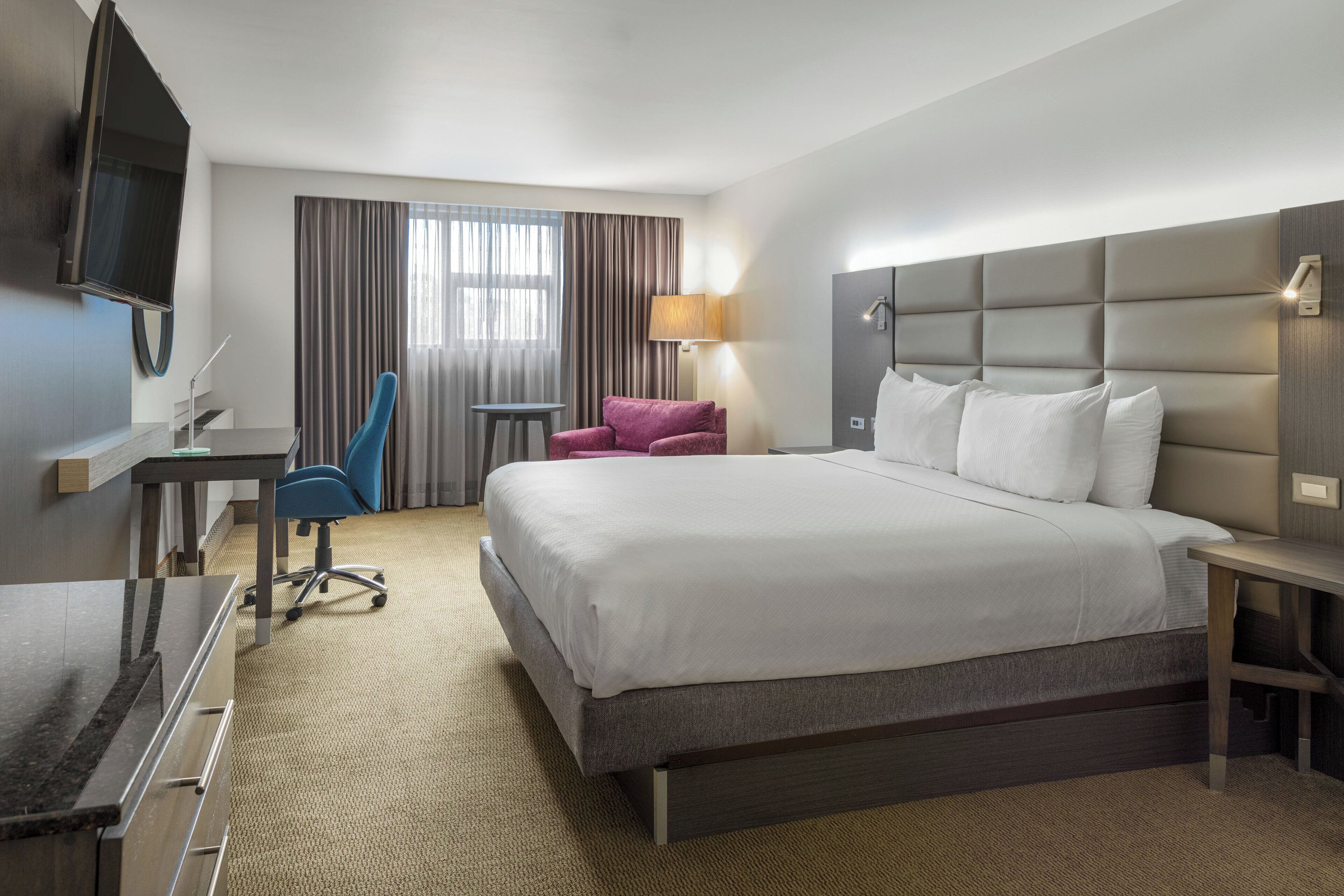 A hotel room with a large bed, a desk with a chair, a TV, and a window with curtains.