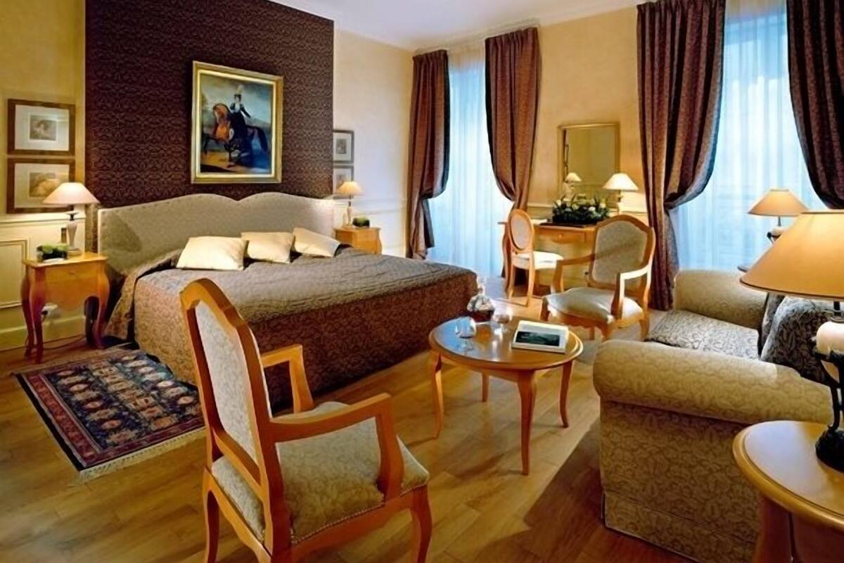 A hotel room with a large bed, two armchairs, a small table, a desk, and a chair. There is a painting on the wall, a window with curtains, and a lamp on the desk.