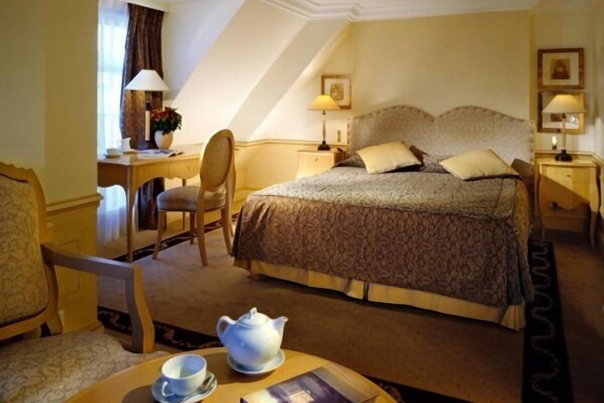 A hotel room with a bed, a desk, a chair, a table with a teapot and a book, and a window with curtains.