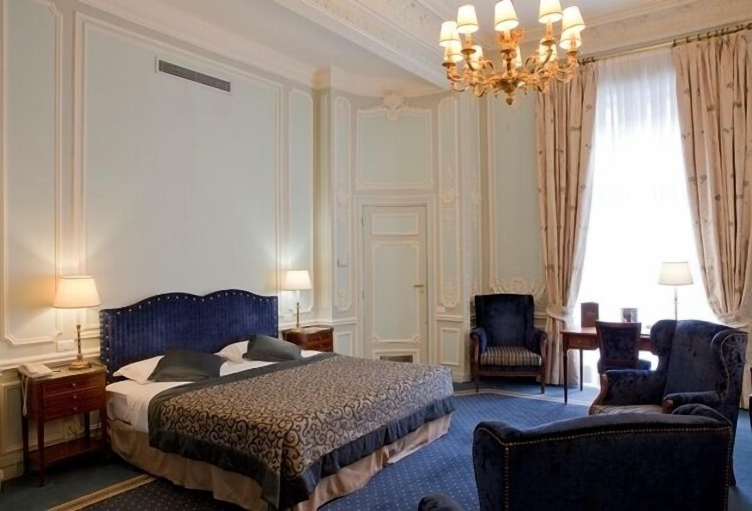 Una camera d'albergo con un letto grande, due comodini, un lampadario, un'area salotto con una poltrona e un tavolino, e una finestra con tende.