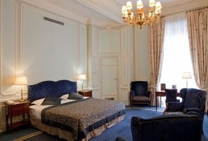A hotel room with a large bed, two nightstands, a chandelier, a sitting area with a chair and a small table, and a window with curtains. - Hotel Lotti (Paris)