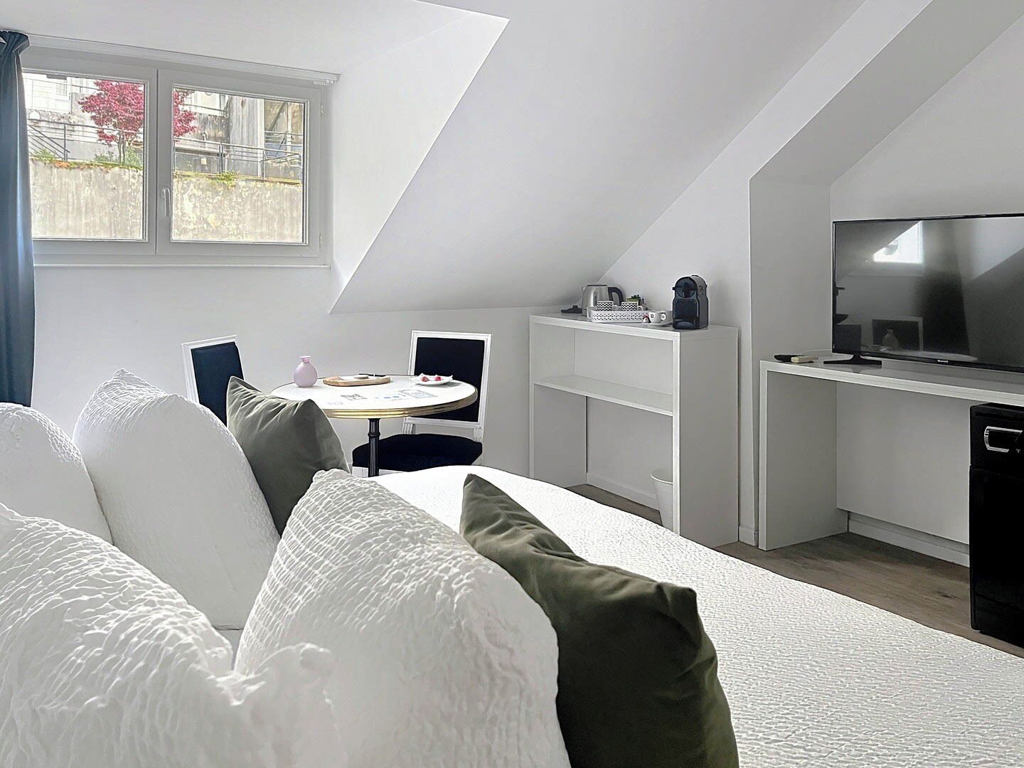 A modern, compact room with a loft-style ceiling, a bed with white and green pillows, a small round table with a pink vase, a television on a stand, and a white cabinet.