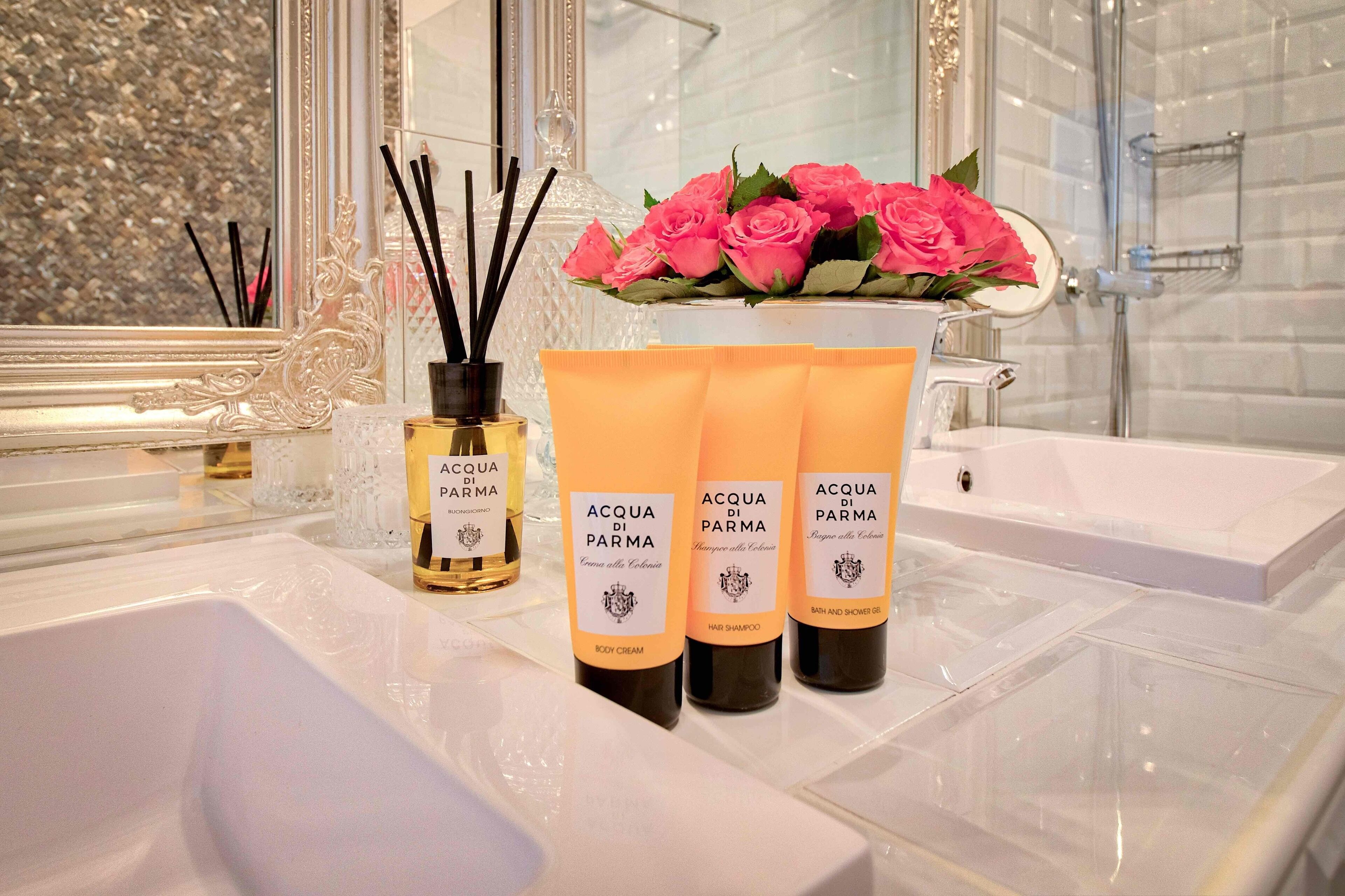 A bathroom with a white sink, a mirror, and three bottles of Acqua di Parma body cream.