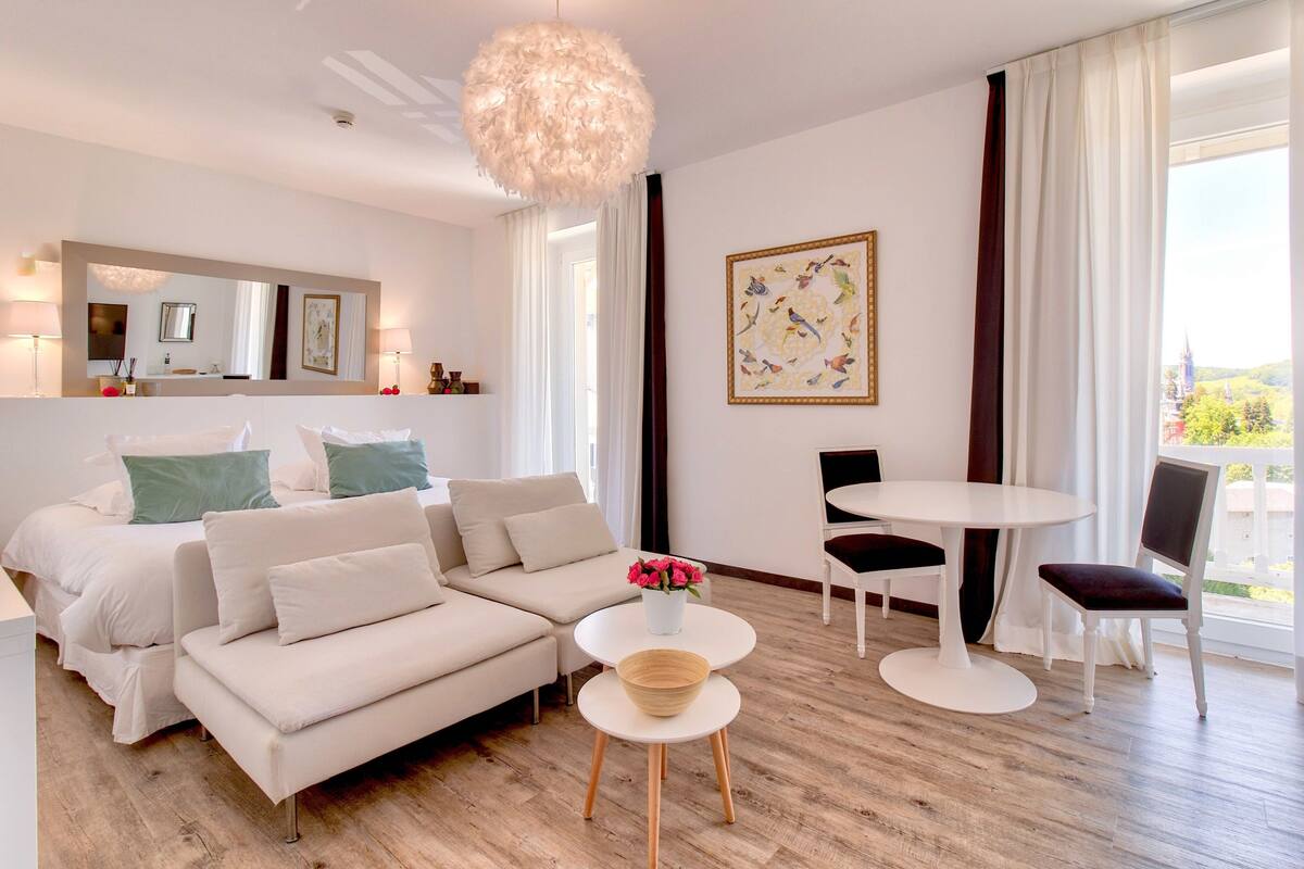 A modern hotel room with a large bed, a sofa, a dining table with chairs, a small side table with a vase of flowers, and a large window with curtains.