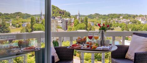 Un balcón con sillas de mimbre, una mesa dispuesta con comida y bebidas, y una vista panorámica de un pueblo con edificios y vegetación.