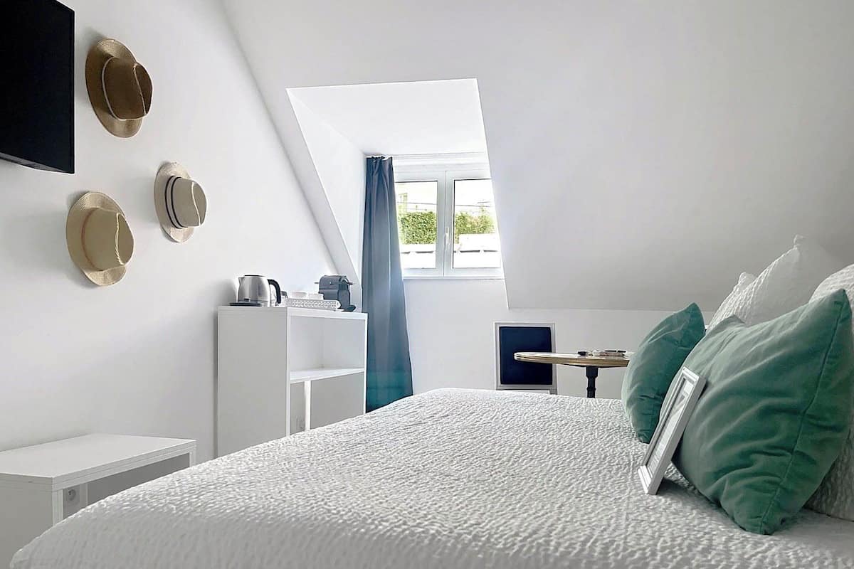 A bedroom with a sloped ceiling, a bed with white bedding, a desk with a computer, and a window with curtains.