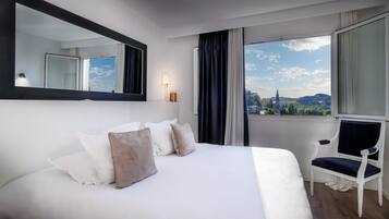 A hotel room with a large bed, a chair, a window with a city view, and a mirror on the wall.
