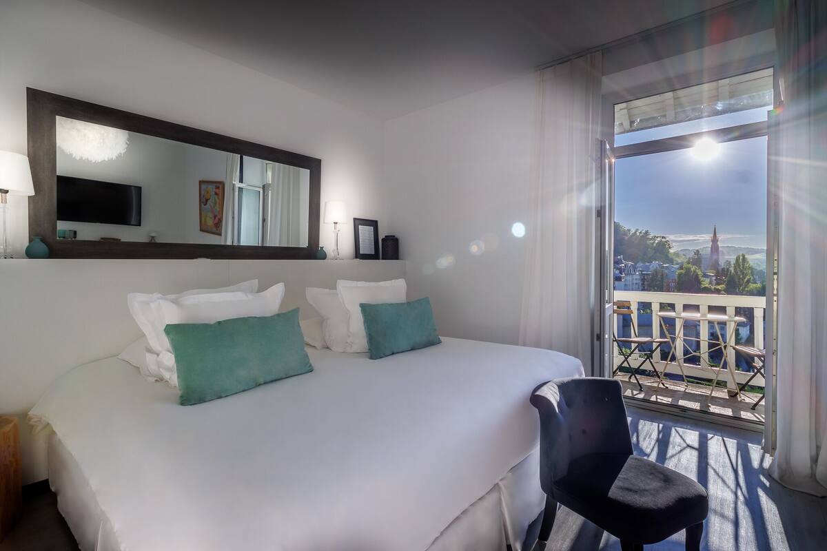 A hotel room with a large bed, a chair, a TV, and a balcony with a view.