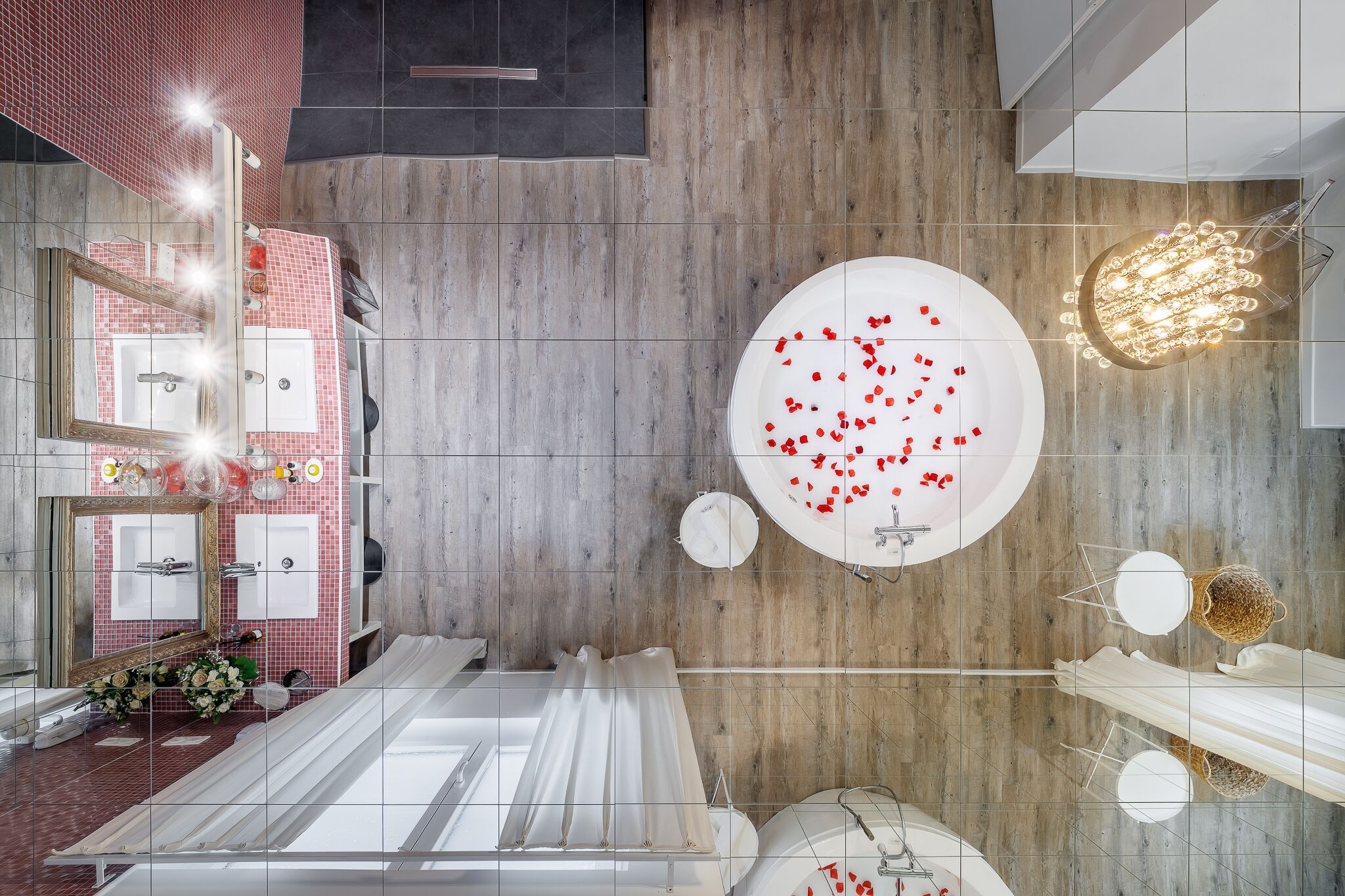 A modern bathroom with a wooden floor, a large mirror, a round sink, and a bathtub with rose petals.