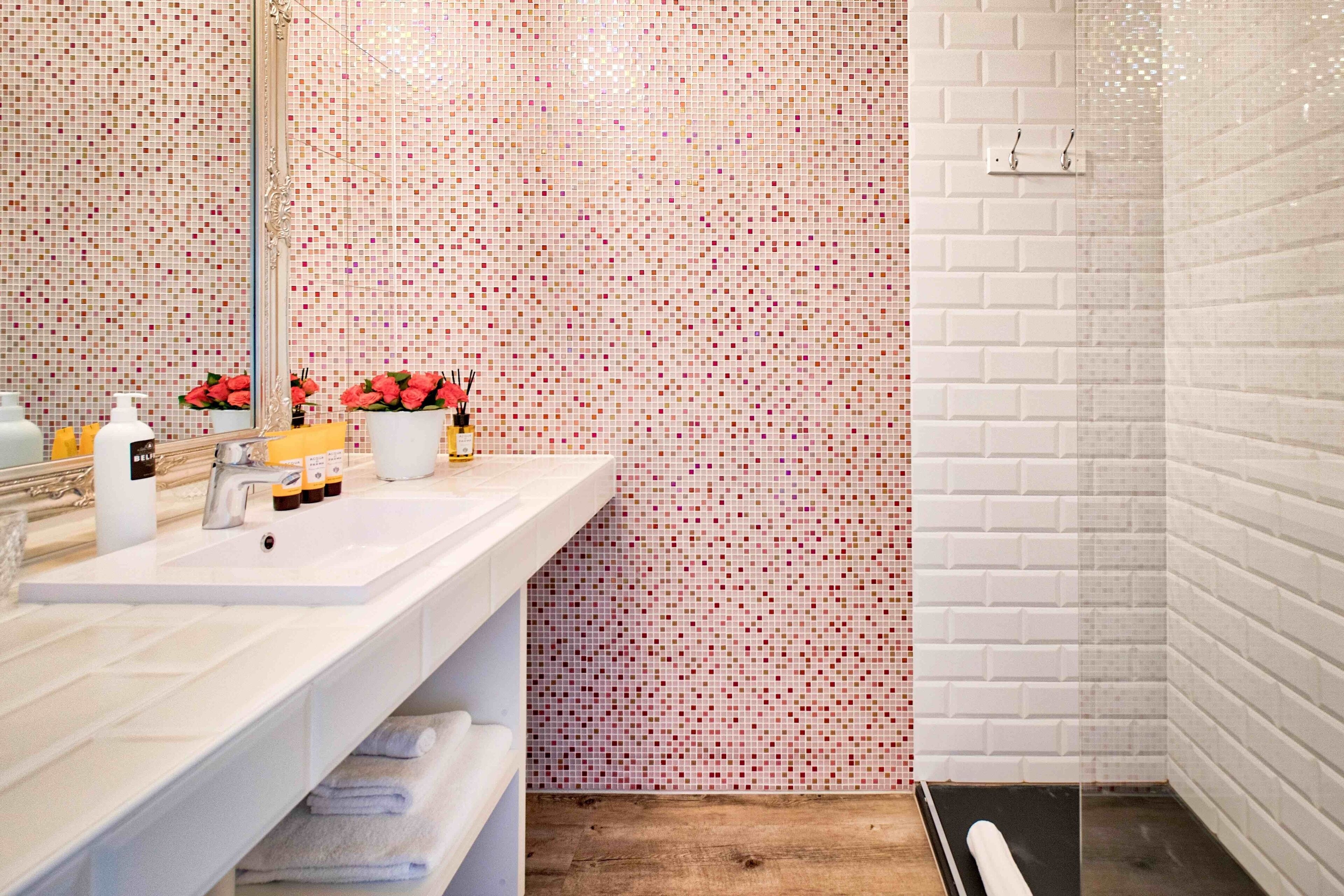 A modern bathroom with a white sink, mirror, and tiled walls. There are toiletries on the sink and a towel rack below.