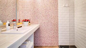 A modern bathroom with a white sink, mirror, and tiled walls. There are toiletries on the sink and a towel rack below.
