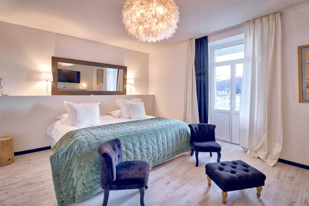 A hotel room with a large bed, two chairs, a small table, a mirror, and a window with curtains.