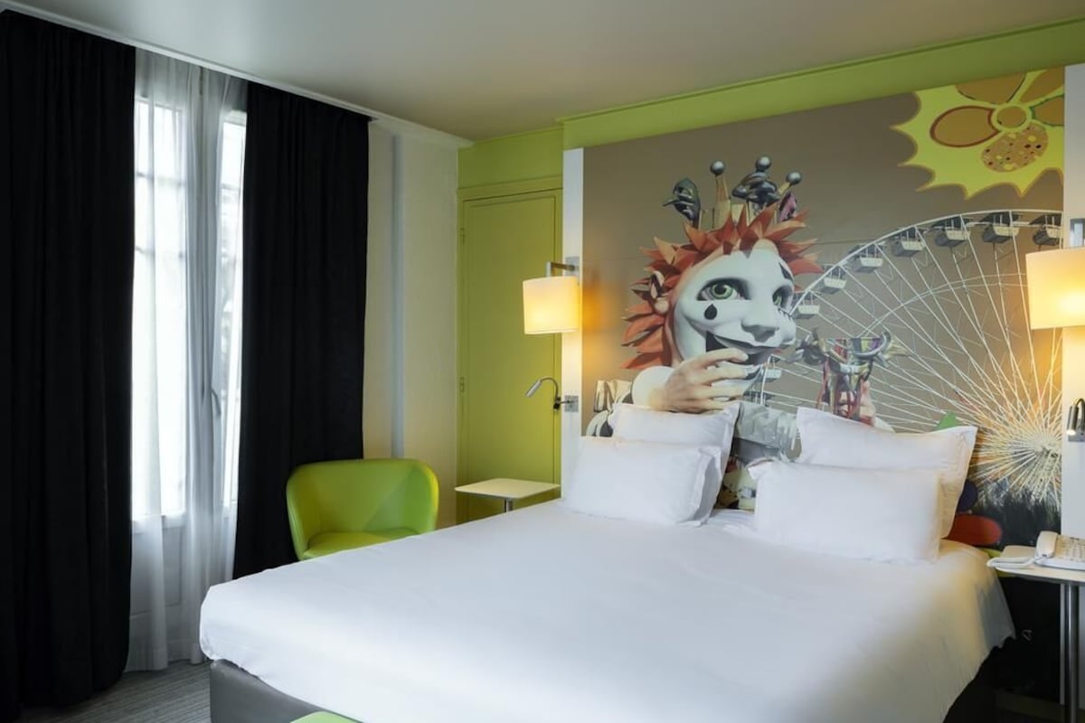 A hotel room with a large bed, a green chair, a desk with a lamp, and a wall mural.