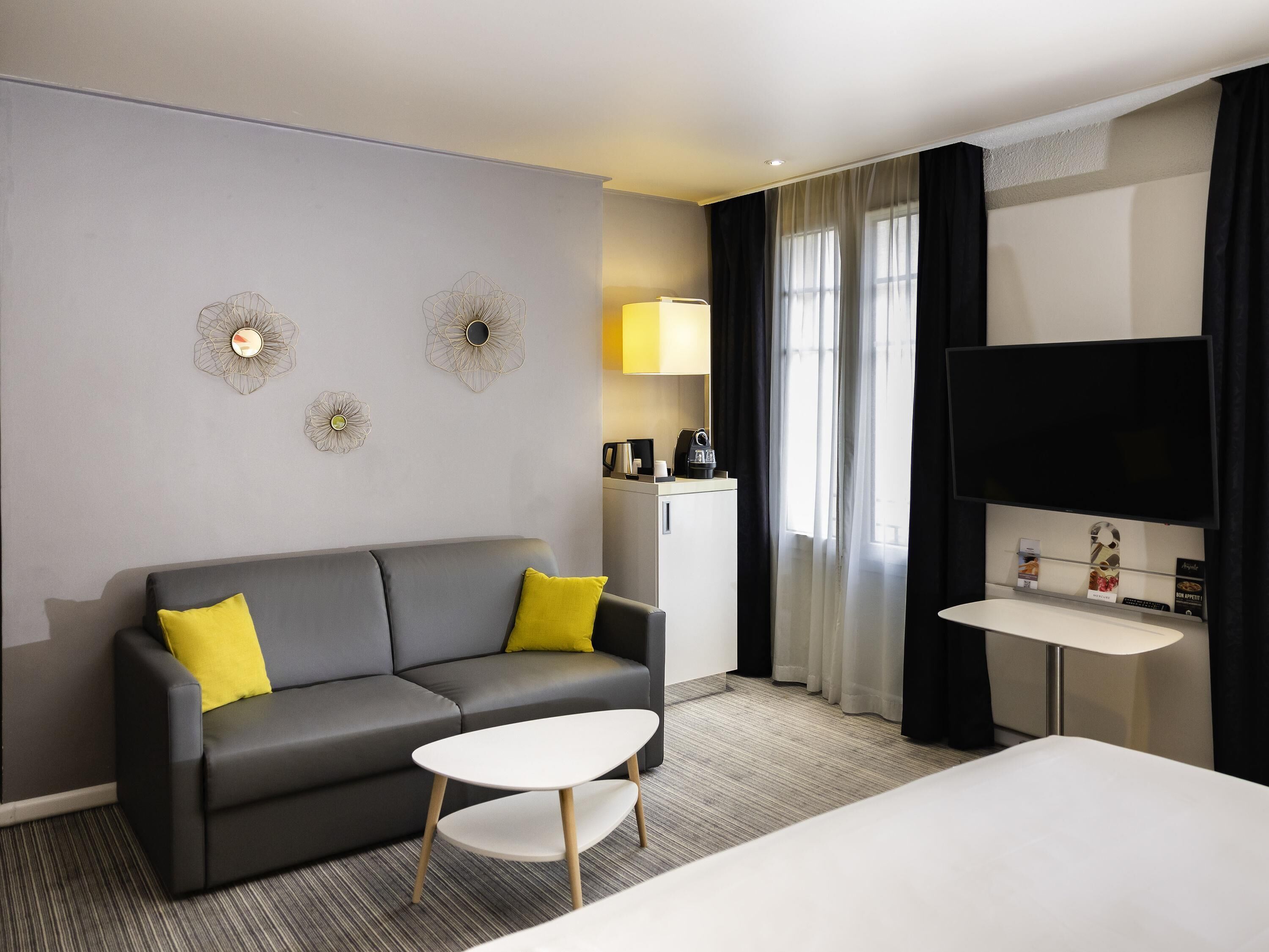 Una habitación de hotel moderna con un sofá gris, una pequeña mesa blanca, una cama, un televisor y un minibar.