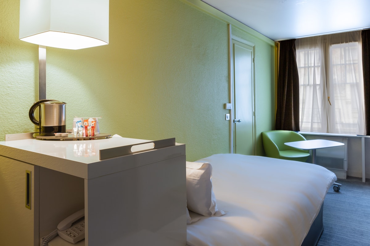 A hotel room with a bed, a desk with a lamp, a chair, and a window with curtains.