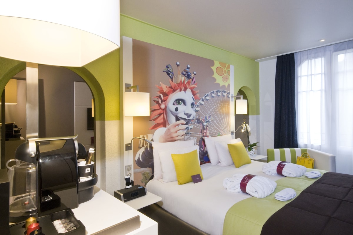 A hotel room with a bed, a coffee machine, a mirror, and a wall with a mural.