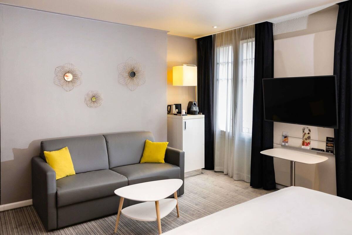 Ein modernes Hotelzimmer mit grauem Sofa, einem kleinen weißen Tisch, einem Bett, einem Fernseher und einer Minibar.