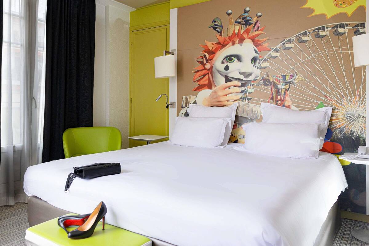 Ein Hotelzimmer mit einem Bett, einem grünen Sessel, einem kleinen Tisch und einer Wand mit Wandmalerei.