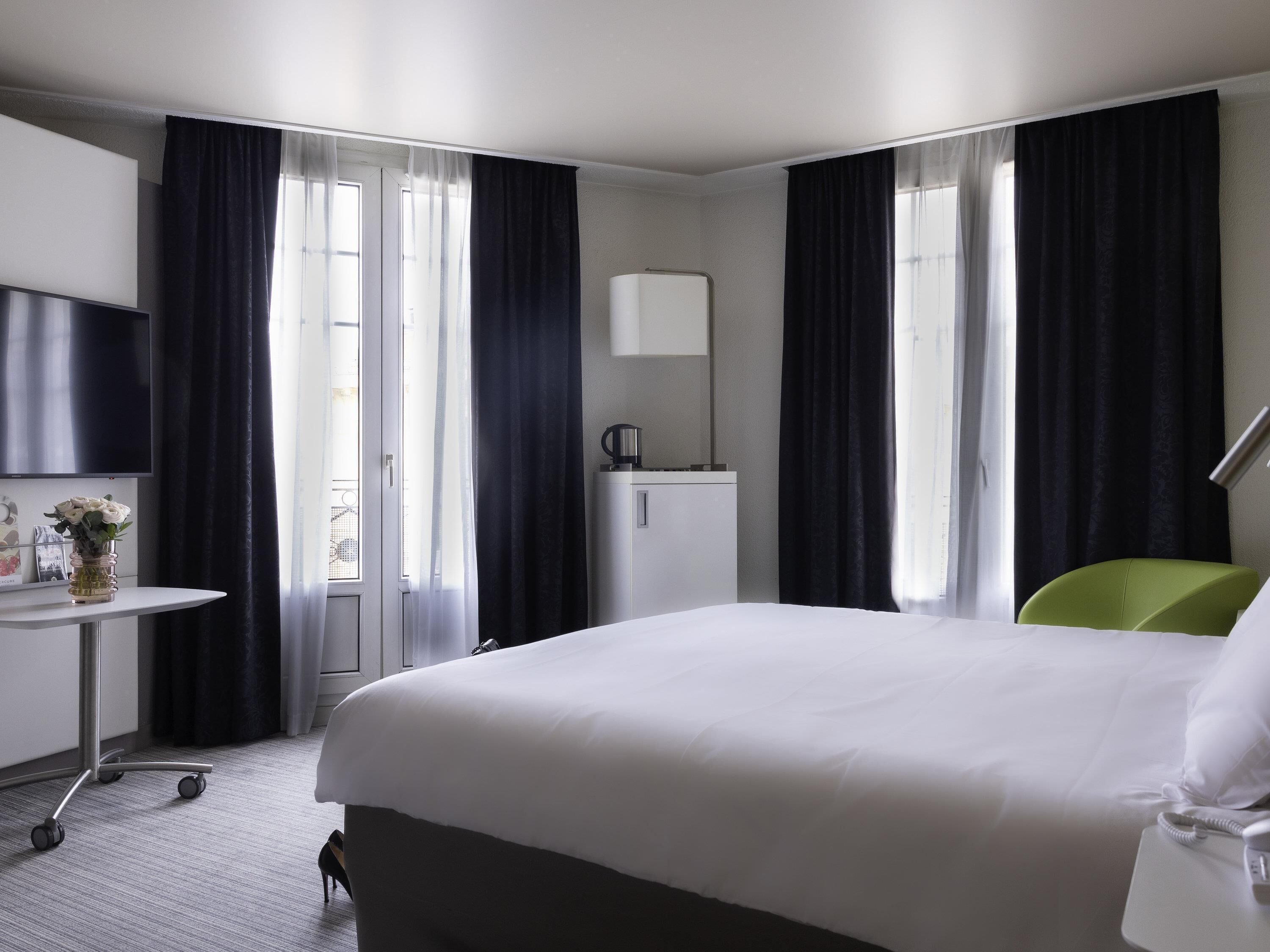 A hotel room with a bed, a desk, a chair, a small table, a lamp, a TV, and a window with curtains.