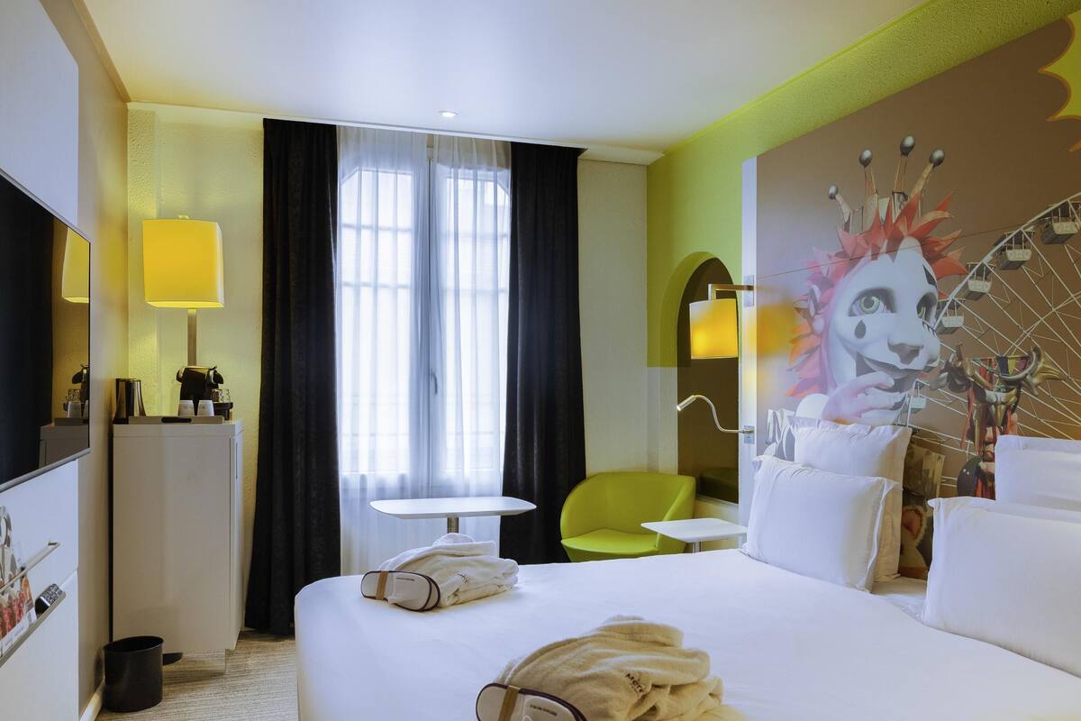 Ein Hotelzimmer mit Bett, kleinem Tisch, Stuhl, Fernseher und einer Wand mit Wandgemälde.